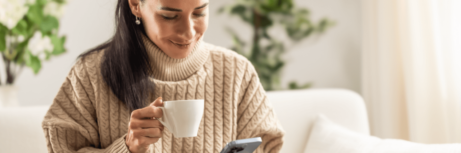 Woman in a chunky jumper holding a mug and happily looking at her phone
